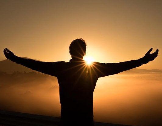A person standing with arms outspread toward the setting sun overlooking clouds probably from on top of a mountain.