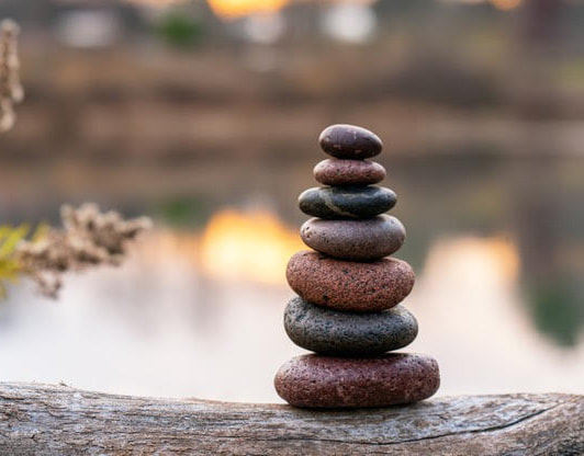 Multi-coloured stoned stacked one on top of the other from smallest at the top to largest on the bottom, balancing on a log by a lake.